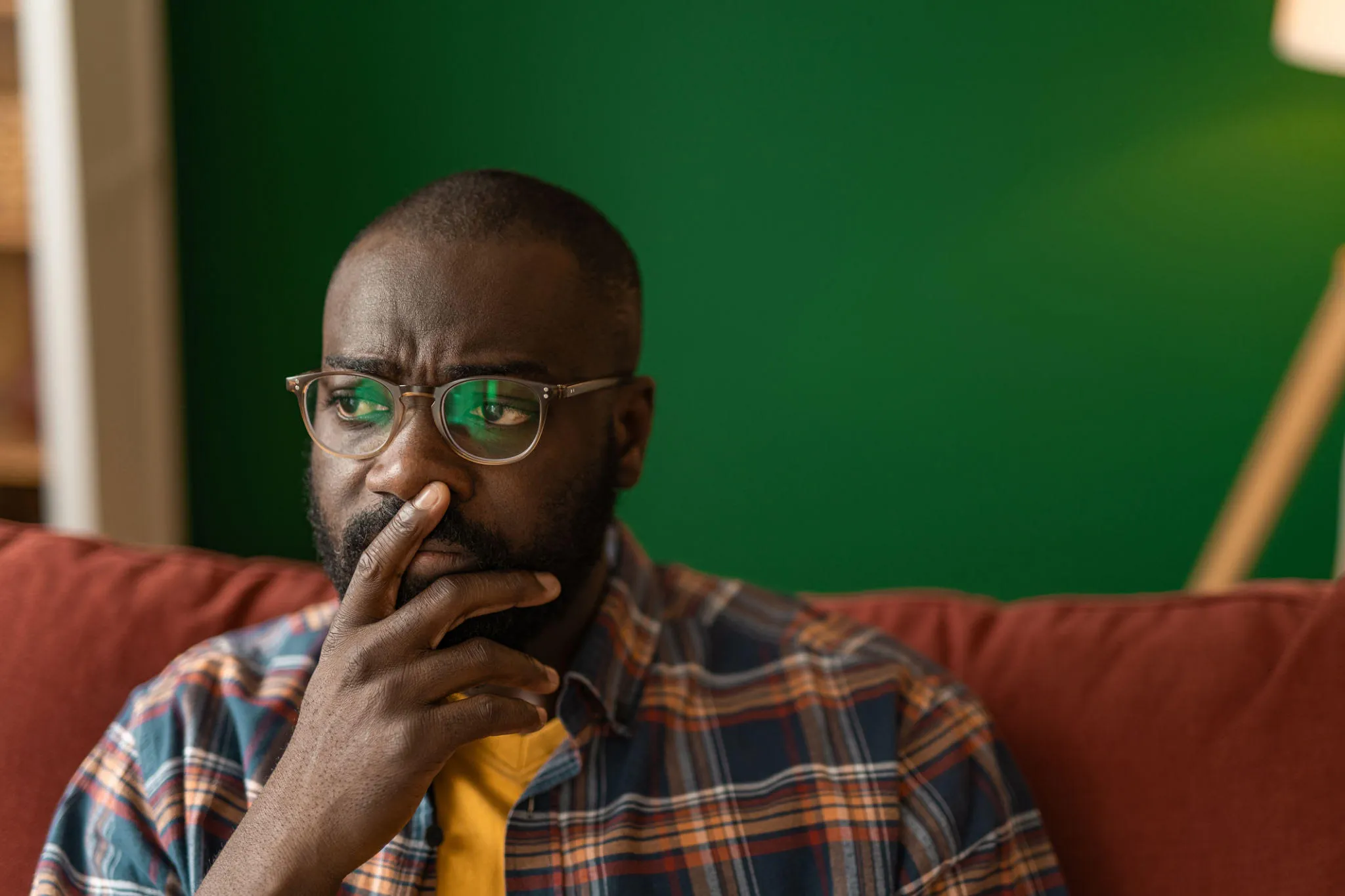 A man sits on a couch, glasses on, thinking something over.